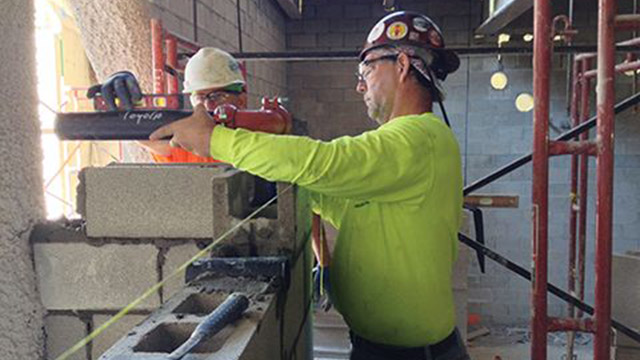 A bricklayer (left) works in alongside a pipefitter (right) to simultaneously construct the block wall and the plumbing contained within it.